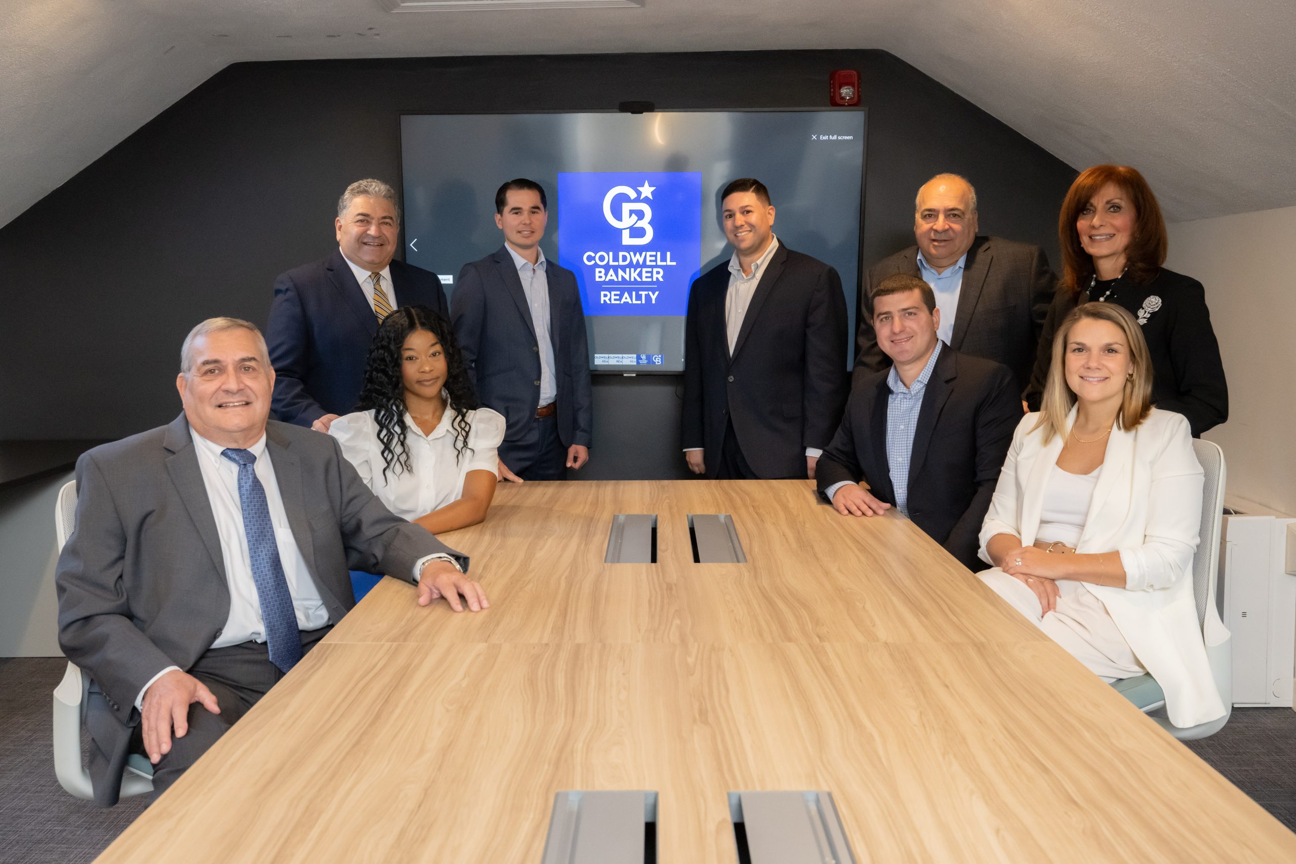 A photo of the DelRose McShane Team posing in the office conference room. The Coldwell banker logo is displayed behind them
