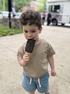 On the Belmont Waltham Line at the Beaver Brook Reservation a boy eats a chocolate eclair ice cream at the park entrance with the ice cream truck in the background