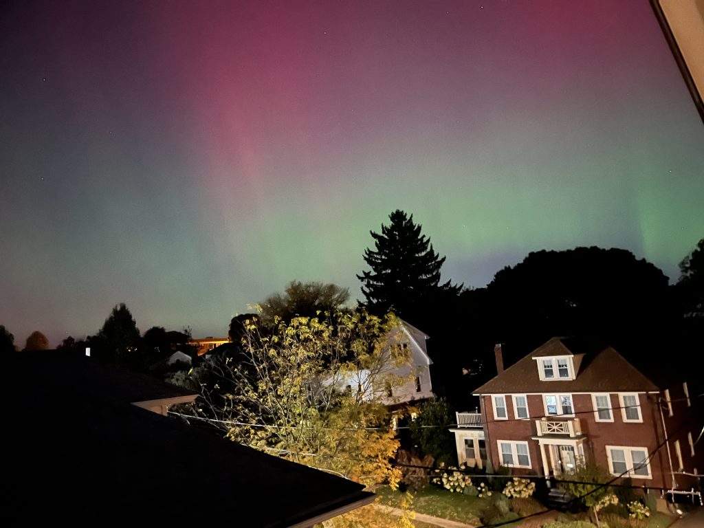 A photo showing Aurora Borealis otherwise known as the Northern Lights. Photo is taken from the third floor of a home, looking over the rooftops of the neighborhood.