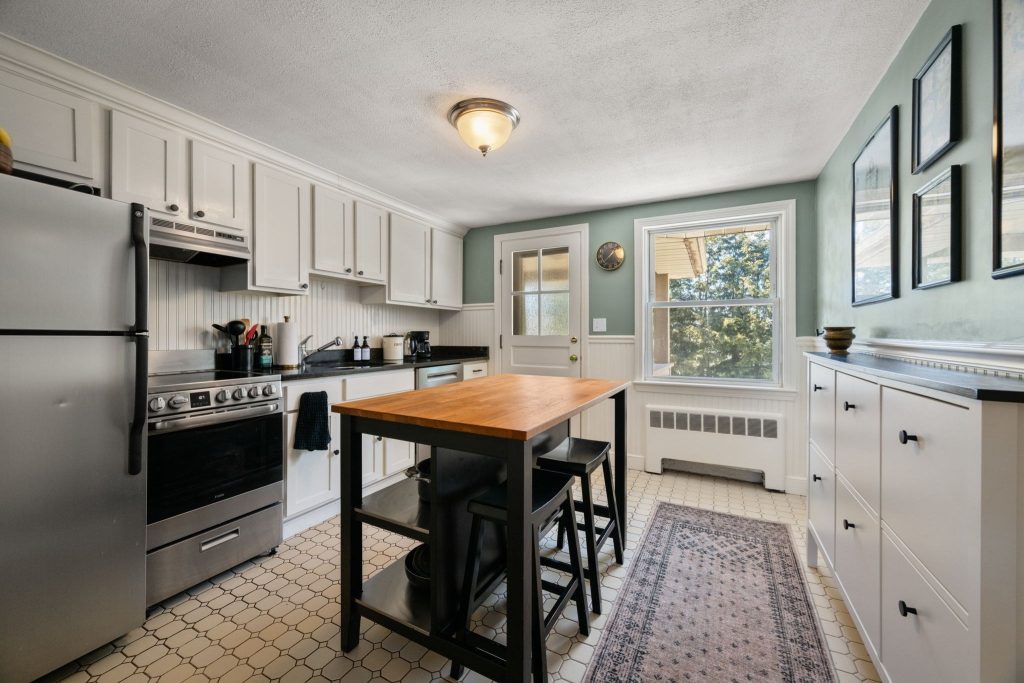 Listing photo of a ktichen with white cabinets and stainless steel appliances. In the center of the photo is a portable kitchen island and seating area, with a carpet to the right. 